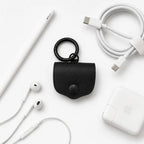 Assomma full-grain leather earphone cases (Ember Natural/Matte Black) with D-ring and round clasp. Flat lay on a white background with a braided cable, Apple Pencil, and earphones, showing a minimalist EDC industrial aesthetic