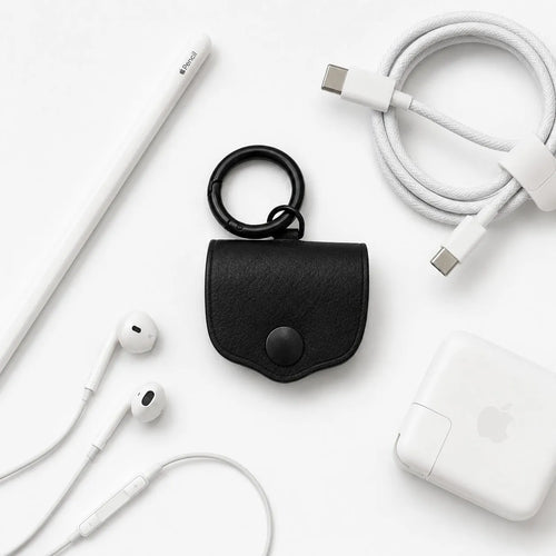 Assomma full-grain leather earphone cases (Ember Natural/Matte Black) with D-ring and round clasp. Flat lay on a white background with a braided cable, Apple Pencil, and earphones, showing a minimalist EDC industrial aesthetic